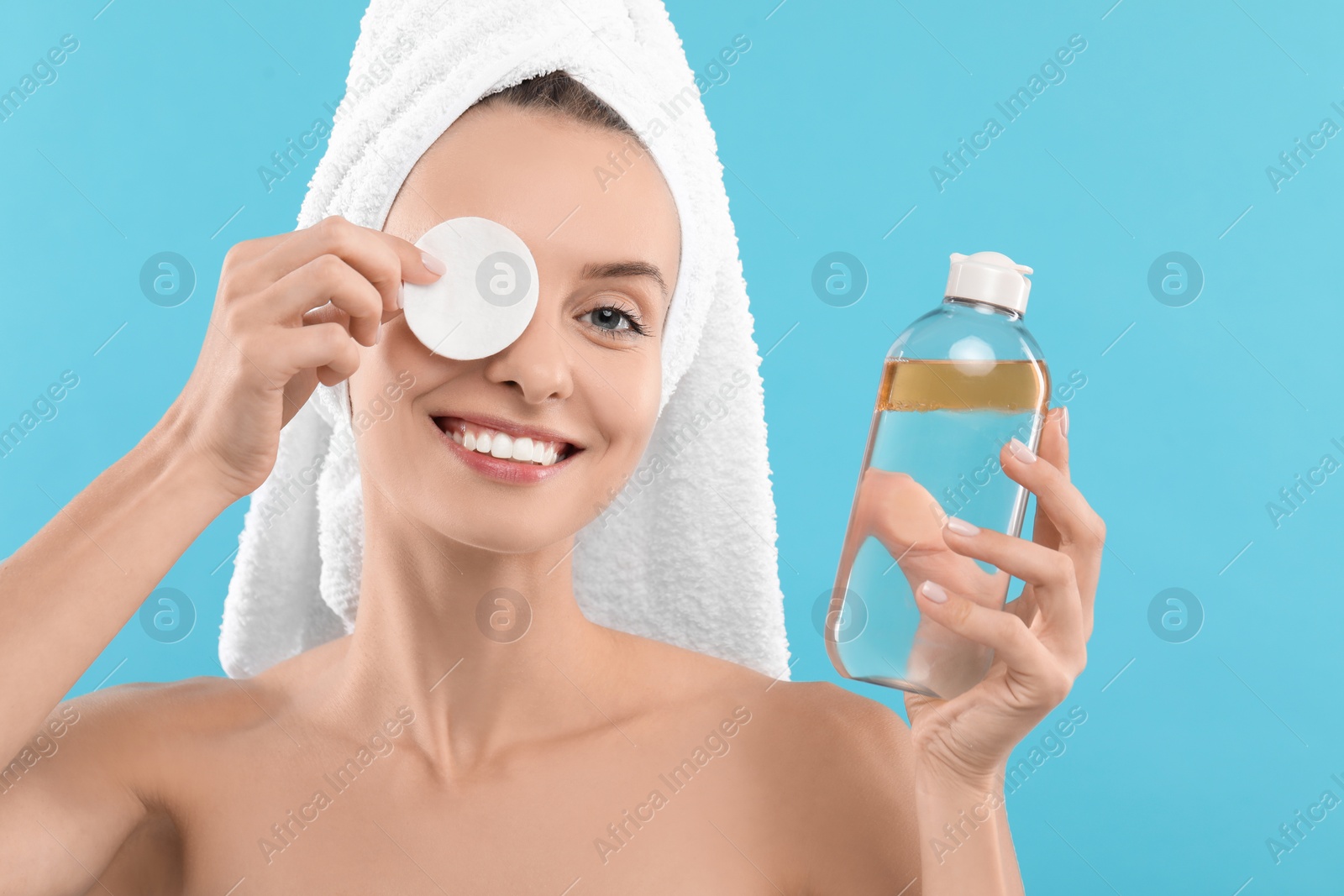 Photo of Smiling woman removing makeup with cotton pad and holding bottle on light blue background