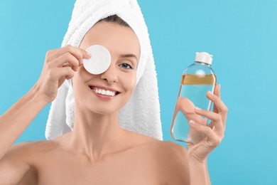 Smiling woman removing makeup with cotton pad and holding bottle on light blue background