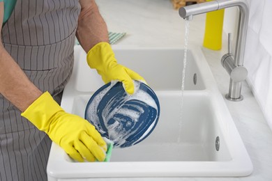 Photo of Man in protective gloves washing plate above sink in kitchen, closeup