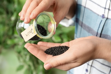 Photo of Woman pouring onion seeds from glass jar into hand against blurred background, closeup. Vegetable planting