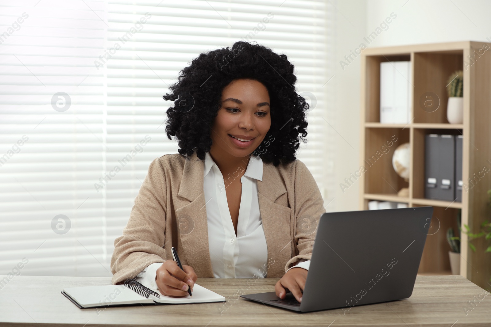 Photo of Happy young woman writing notes while using laptop at wooden desk indoors