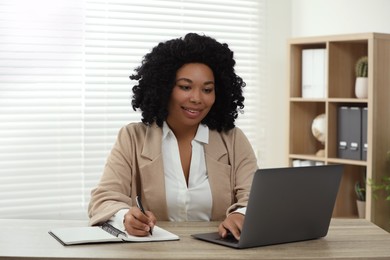 Happy young woman writing notes while using laptop at wooden desk indoors