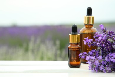 Bottles of essential oil and lavender flowers on white wooden table in field, space for text