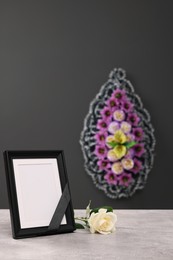 Photo frame with black ribbon, rose on light table and wreath of flowers near grey wall indoors, space for text. Funeral attributes