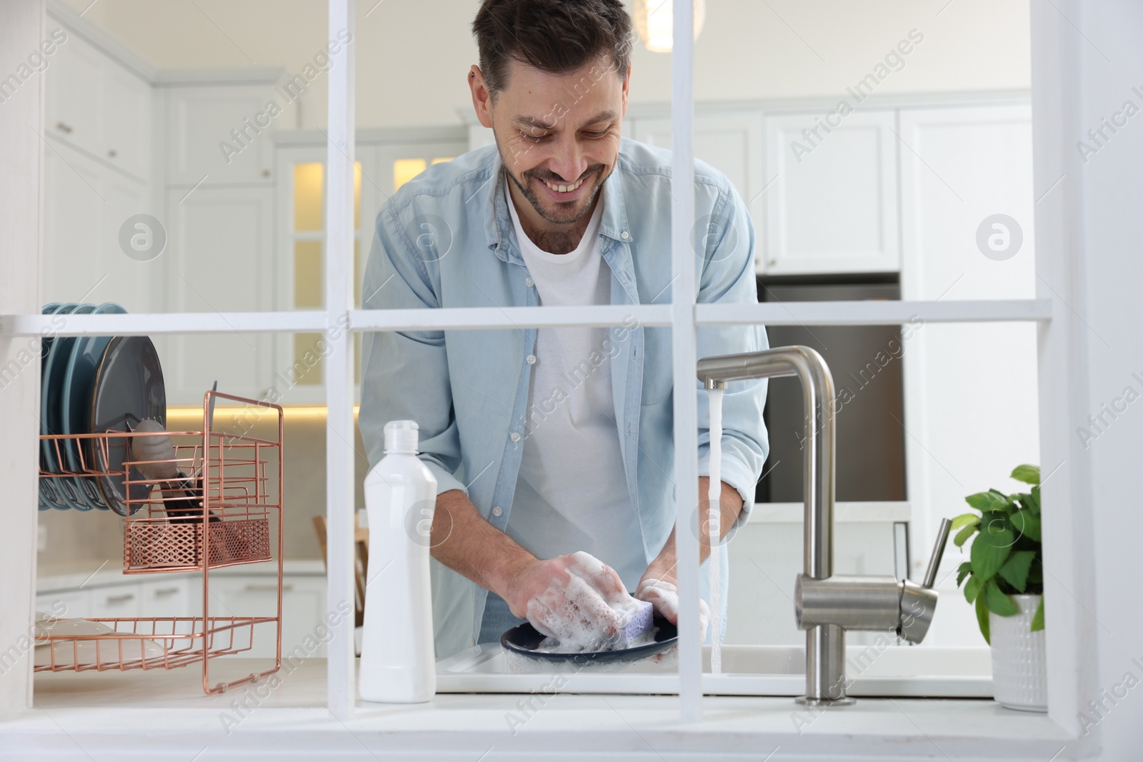 Photo of Man washing plate above sink in kitchen, view from outside