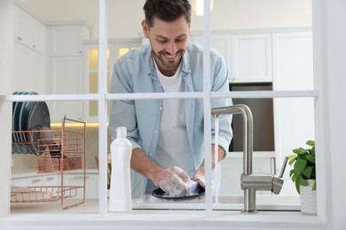 Photo of Man washing plate above sink in kitchen, view from outside