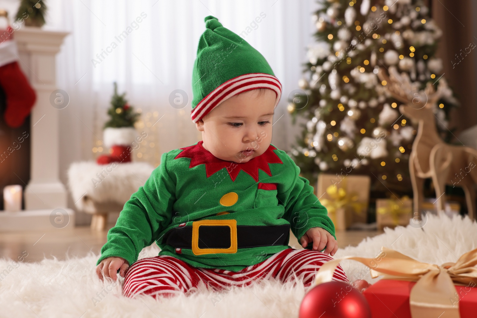 Photo of Baby wearing cute elf costume on floor in room decorated for Christmas