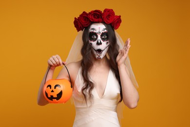 Photo of Emotional young woman in scary bride costume with sugar skull makeup, flower crown and pumpkin bucket on orange background. Halloween celebration