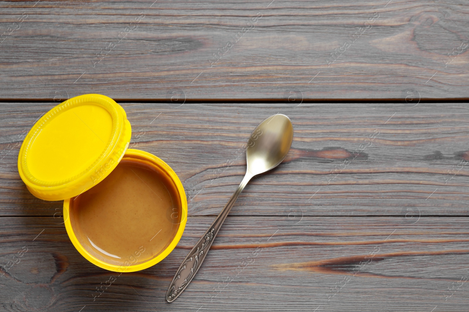 Photo of Spicy mustard in plastic container and spoon on grey wooden table, flat lay