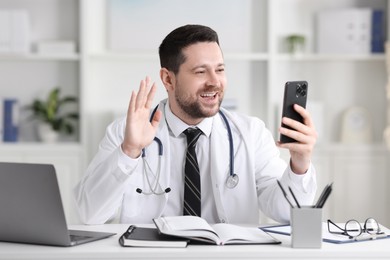 Smiling doctor having online consultation via smartphone at table in clinic