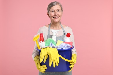 Photo of Happy housewife holding bucket with cleaning supplies on pink background