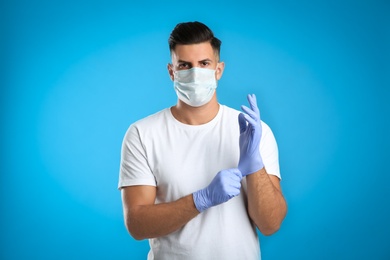 Man in protective face mask putting on medical gloves against blue background