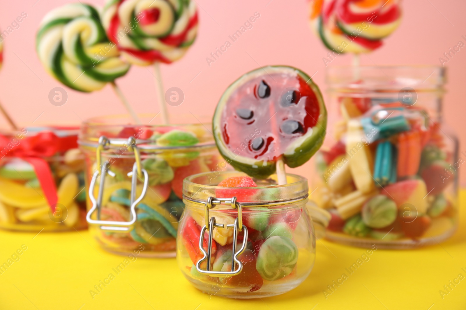 Photo of Tasty colorful candies in glass jars on yellow table against pink background