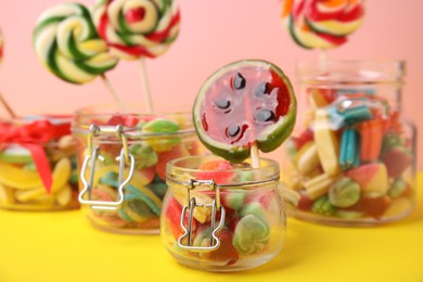 Tasty colorful candies in glass jars on yellow table against pink background