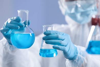 Scientist holding flasks with light blue liquid on grey background, closeup