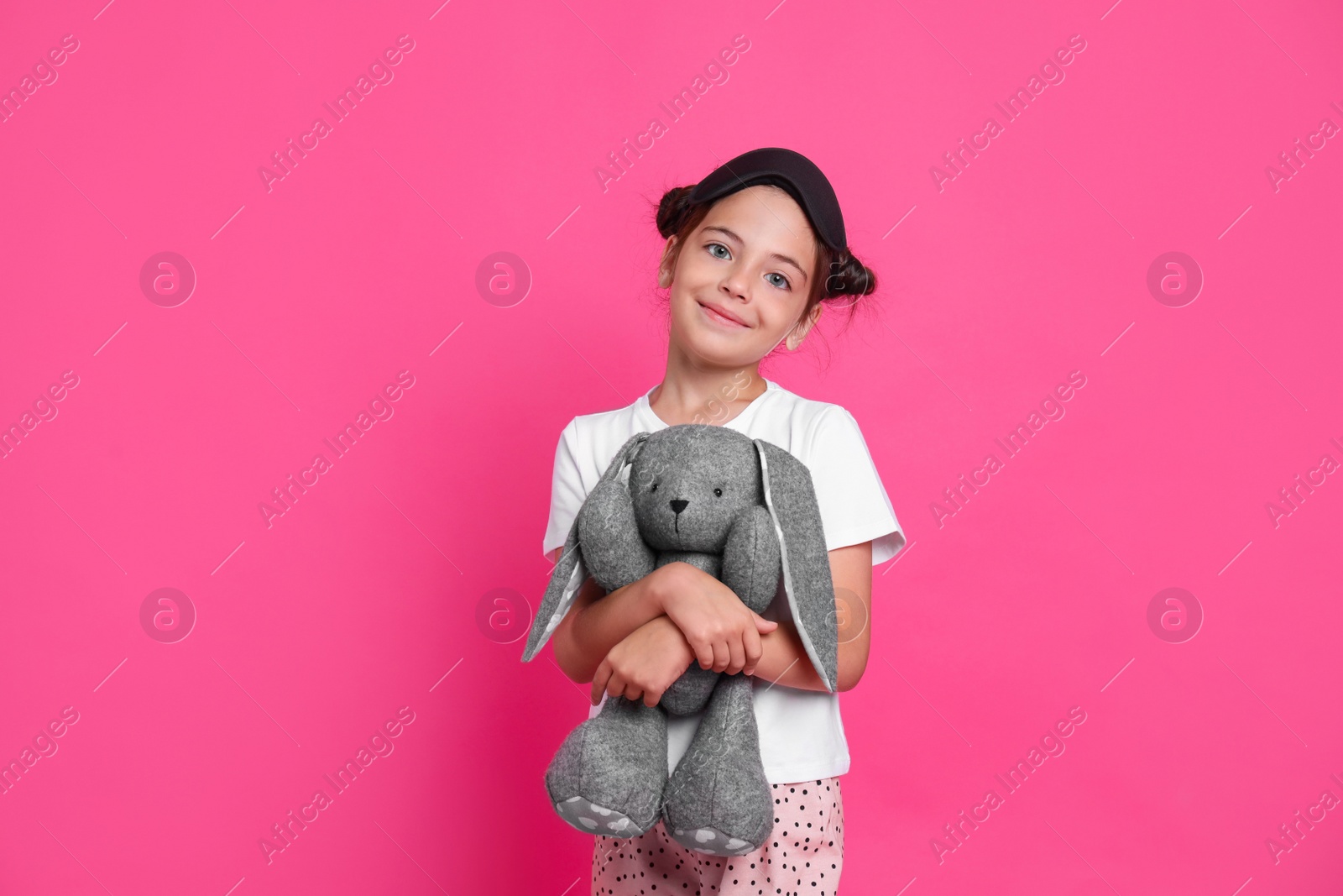 Photo of Cute girl in pajamas with sleep mask and toy bunny on pink background