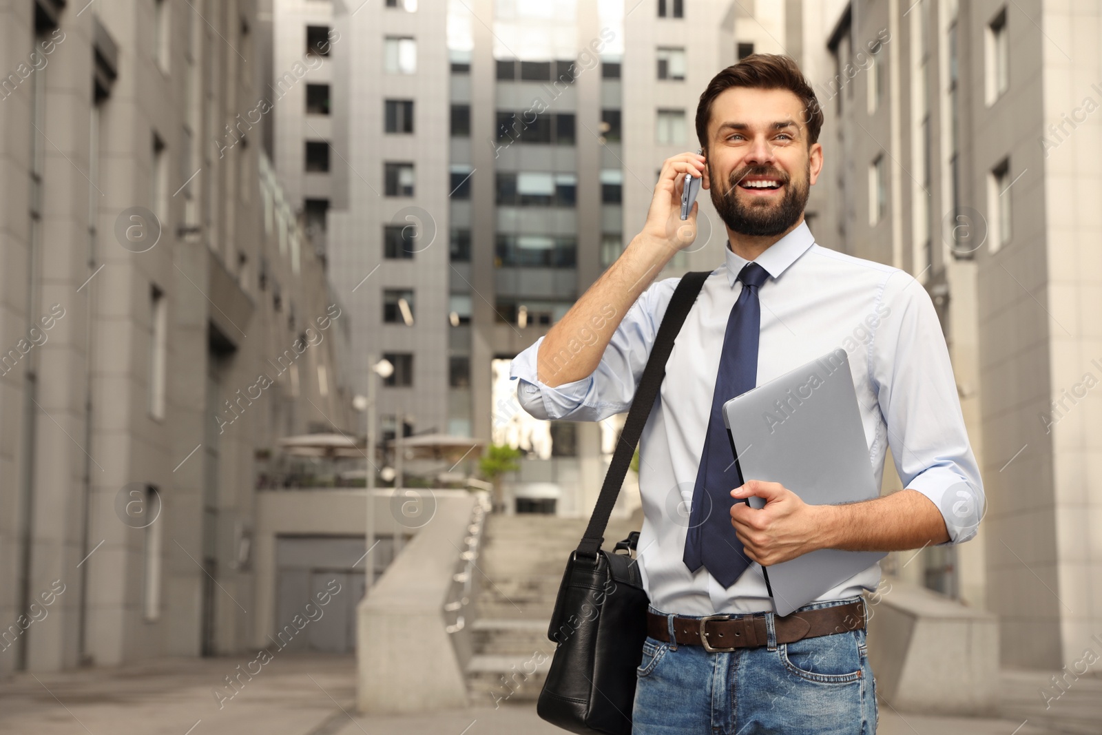 Photo of Handsome man with laptop talking on phone in city