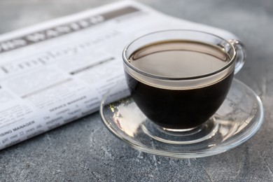 Glass cup with coffee and newspaper on grey table, closeup. Morning ritual