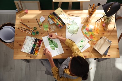 Photo of Young woman drawing leaf with watercolors at table indoors, top view