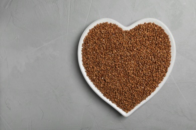 Photo of Heart shaped plate with raw buckwheat on grey background, top view. Space for text