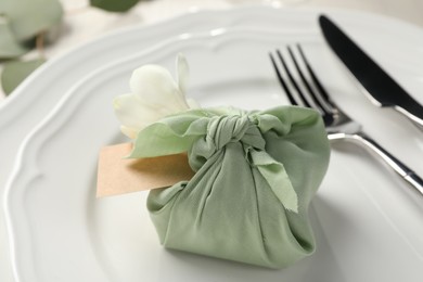 Photo of Furoshiki technique. Gift packed in green fabric with flower and blank card on plate, closeup