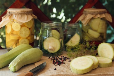 Photo of Cut fresh zucchini and jars of pickled vegetables on white wooden table