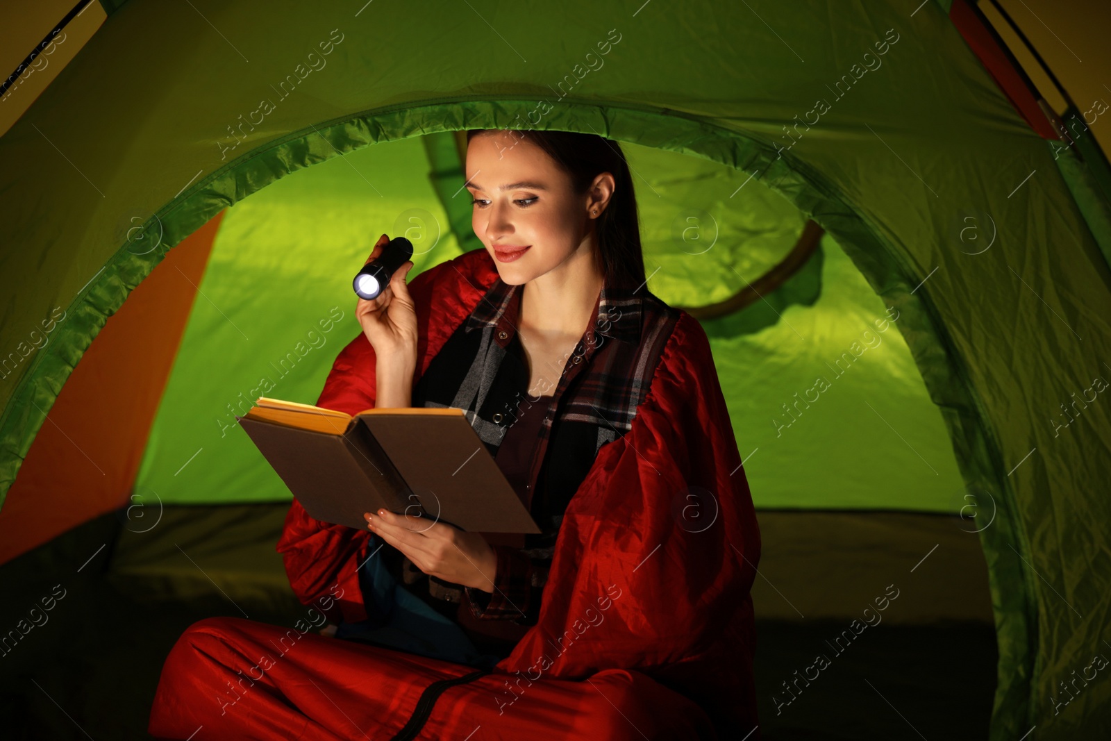 Photo of Young woman with flashlight reading book in tent