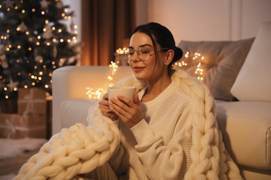 Photo of Young woman with cup of hot drink near sofa at home. Christmas celebration