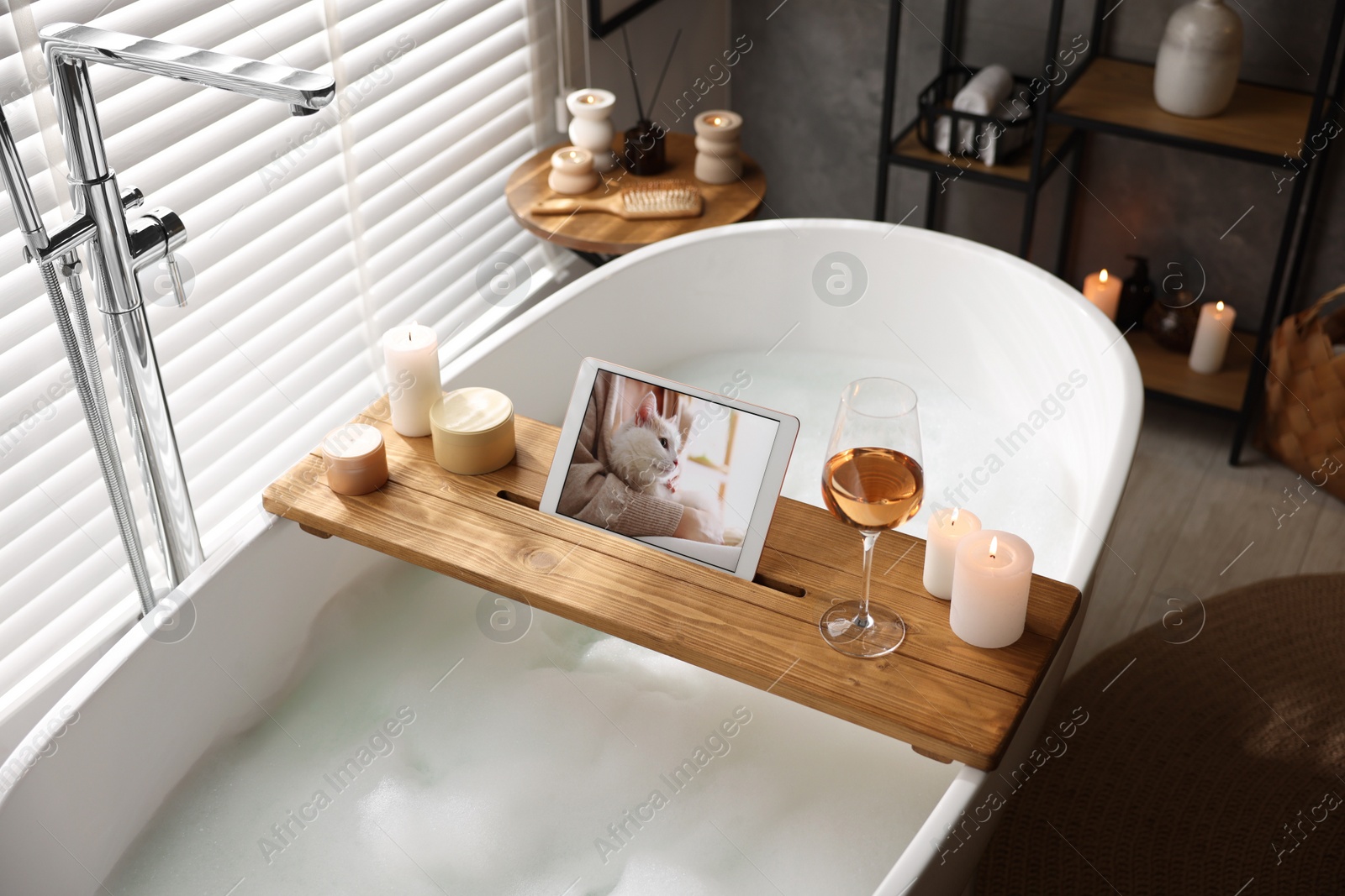 Photo of Wooden tray with tablet, wine and candles on bathtub in bathroom