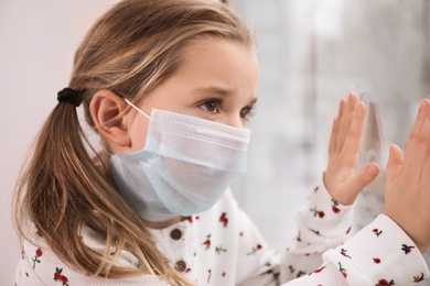 Photo of Sad little girl in protective mask looking out of window indoors. Staying at home during coronavirus pandemic