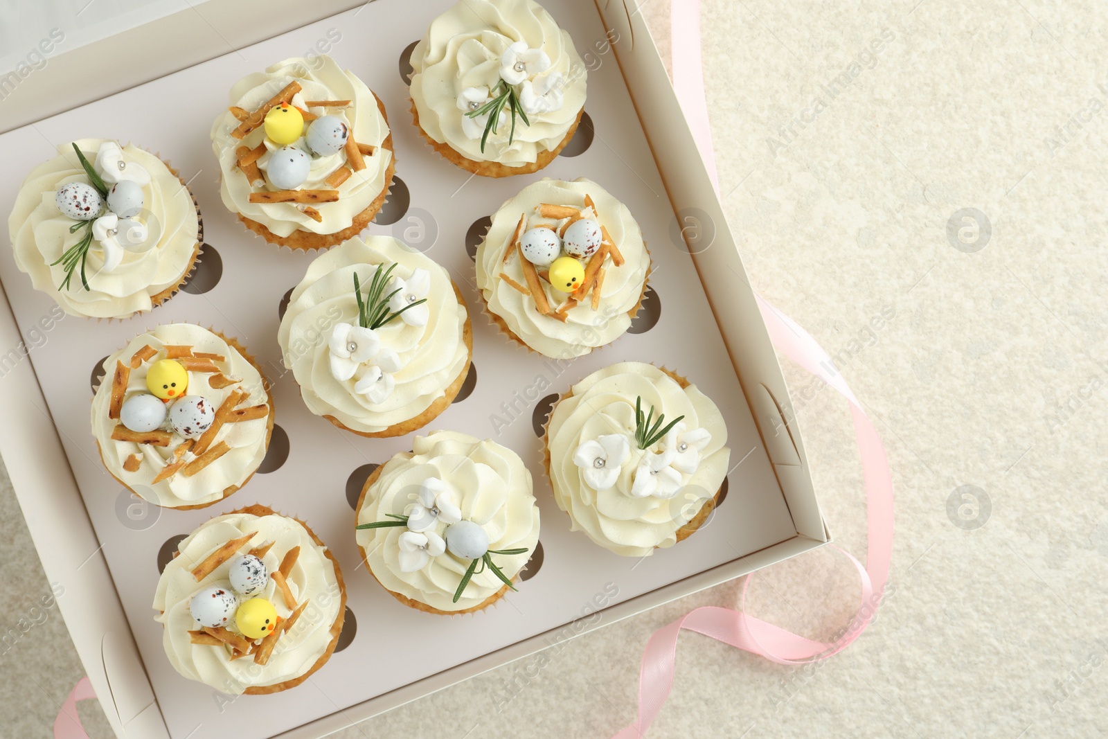 Photo of Tasty Easter cupcakes with vanilla cream in box and pink ribbon on beige table, flat lay. Space for text