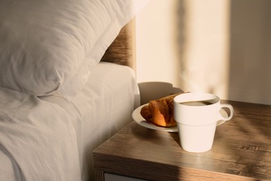 Photo of Cup of coffee and croissant on night stand near bed in morning