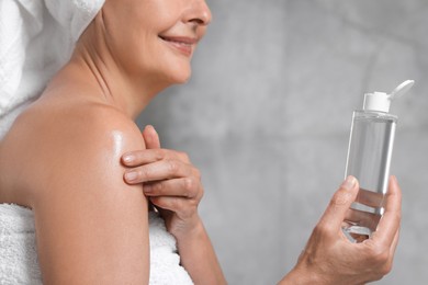 Happy woman applying body oil onto shoulder near grey wall, closeup