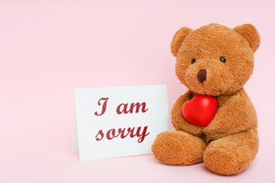 Cute teddy bear holding red heart near card with phrase I Am Sorry on pink background, space for text