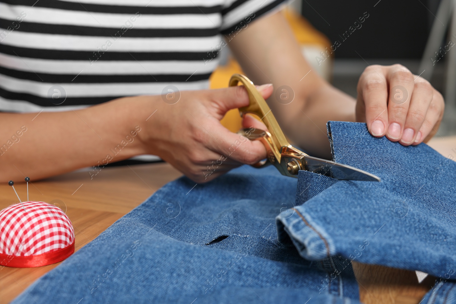 Photo of Young woman shortening jeans with scissors at wooden table, closeup