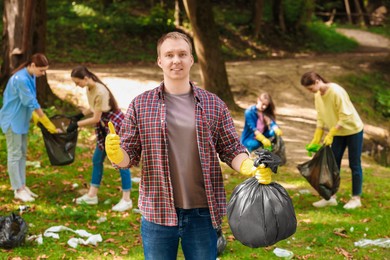Photo of Young man with plastic bag showing thumb up and group of people collecting garbage in park