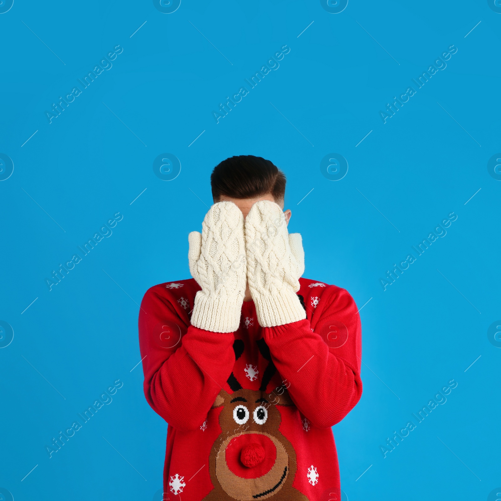 Photo of Man in Christmas sweater and mittens hiding his face on blue background