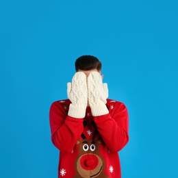 Photo of Man in Christmas sweater and mittens hiding his face on blue background