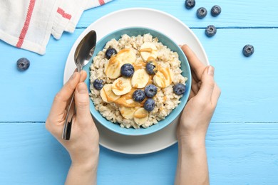 Photo of Woman eating tasty oatmeal with banana, blueberries and peanut butter at light blue wooden table, top view