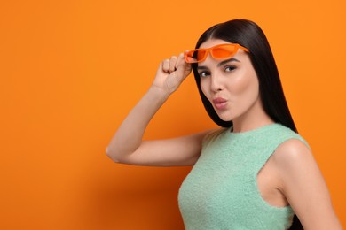 Beautiful young woman in stylish sunglasses blowing kiss on orange background. Space for text