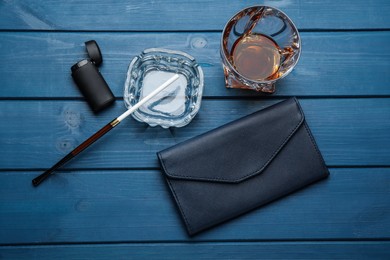 Ashtray with long cigarettes holder, lighter, clutch and alcohol drink on blue wooden table, flat lay