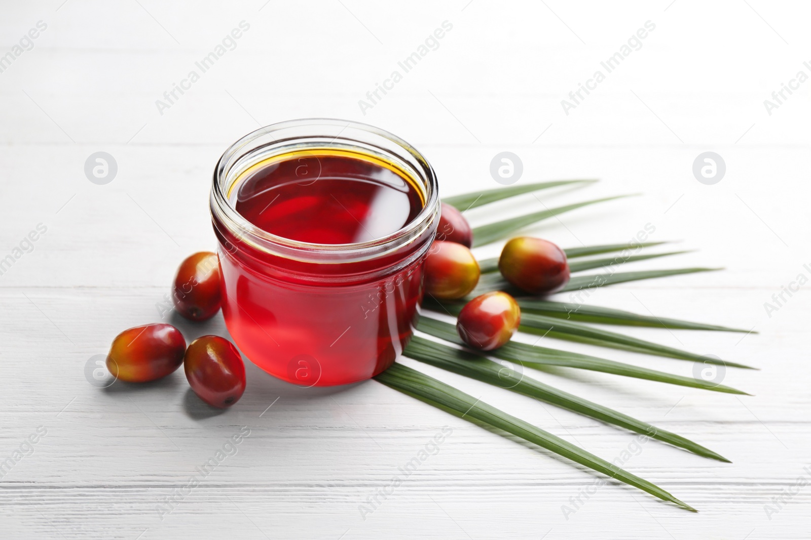 Photo of Palm oil in glass jar, tropical leaf and fruits on white wooden table