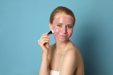 Young woman applying pomegranate face mask on light blue background