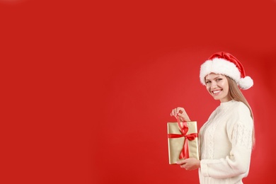 Happy young woman in Santa hat with Christmas gift on red background. Space for text