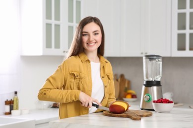 Woman preparing mango for tasty smoothie at white marble table in kitchen