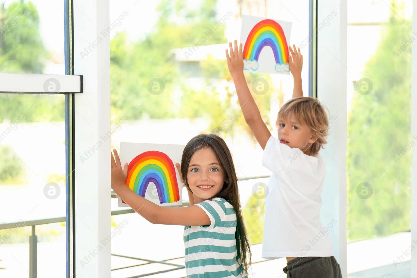 Photo of Little children holding rainbow paintings near window indoors. Stay at home concept