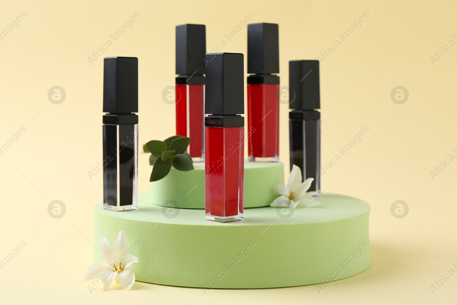 Photo of Different lip glosses, podiums and flowers on beige background