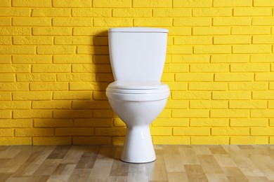 Photo of New clean toilet bowl near yellow brick wall indoors. Interior design