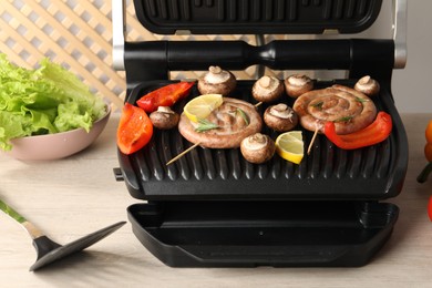 Photo of Electric grill with homemade sausages, bell pepper and mushrooms on wooden table