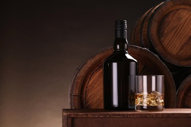 Whiskey with ice cubes in glass and bottle on wooden table near barrels against dark background, space for text